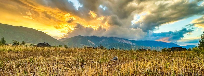 A summer evening looking west across Patio Springs toward the Wasatch — golden grass in the foreground, storm clouds catching the last light of sunset.